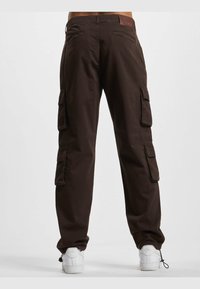 Pantalones cargo marrones hechos de una tela resistente, con múltiples bolsillos laterales y un parche de logo en la cinturilla. Zapatillas blancas complementan el atuendo.