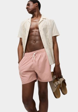 Costumini da bagno rosa corti con vita elastica. Il modello indossa una camicia beige ricamata a maniche corte e tiene in mano dei sandali color cammello.