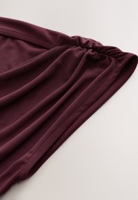 Gros plan sur un tissu bordeaux foncé avec un détail froncé élastique et une couture nette sur un fond clair.