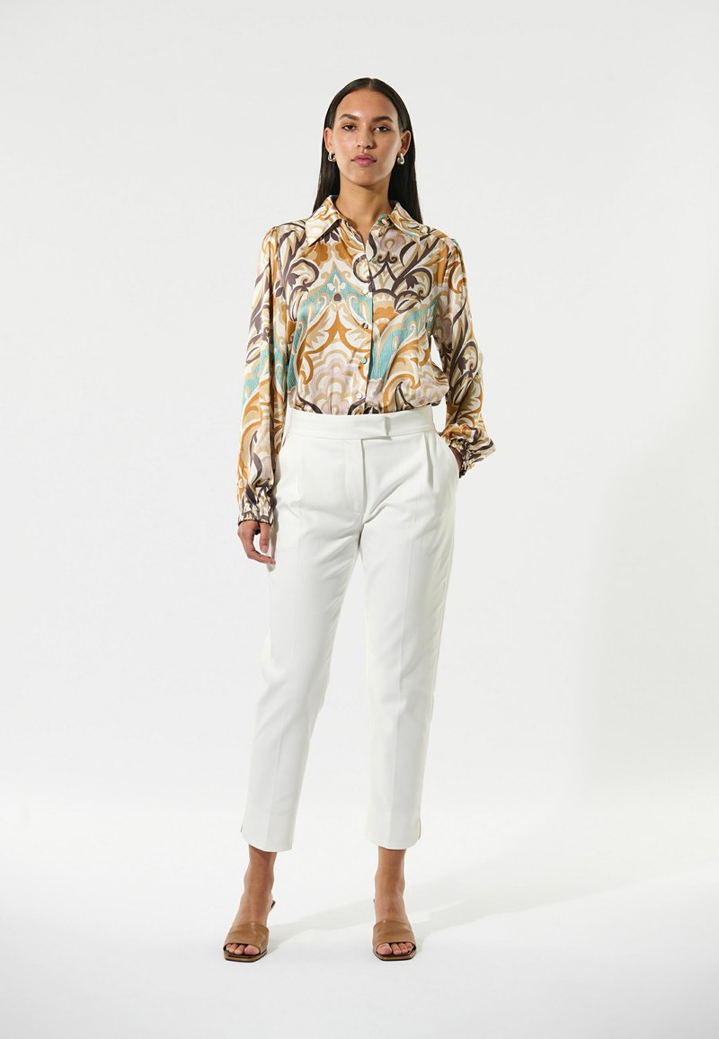 Blouse à manches longues avec un motif élaboré dans des couleurs chaudes, associée à un pantalon blanc bien coupé et des sandales beiges, présentant une coupe relaxée et une texture lisse.