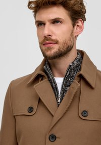 Brauner Wollmischungsmantel mit Reverskragen, doppelter Knopfniederlage und schwarzen Knöpfen, über einem strukturierten grauen Zip-By-Pullover geschichtet.