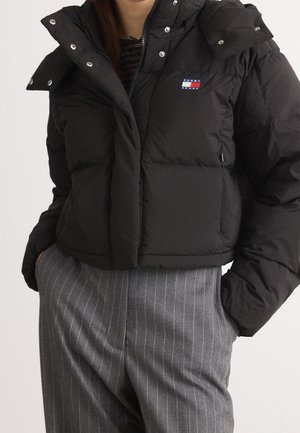 Schwarze, kurz geschnittene Steppjacke mit Tommy Jeans-Logo auf der Brust, getragen zu hoch taillierten grauen Nadelstreifenhosen, Hände in den Taschen.