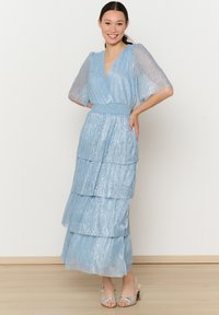 Jasnoniebieska, warstwowa maxi sukienka o połyskującej teksturze, z dekoltem w serek i przezroczystymi, bufkowymi rękawami. Noszona z sandałami na paski w kolorze srebrnym.