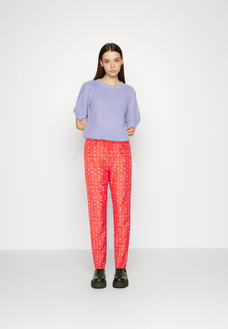 Mujer de pie contra un fondo blanco, vistiendo un suéter corto de manga en color lila, pantalones de estampado naranja brillante y botas de plataforma negras.