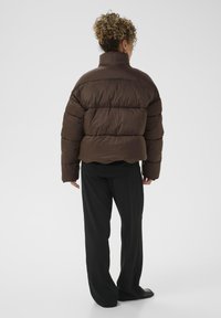 Rudas pūkuotasis švarkas su aukštu koloriumi, suklijuota tekstūra ir kvadratine forma. Dėvimas ant juodos aprangos, derinamas su laisvais juodais kelnais.