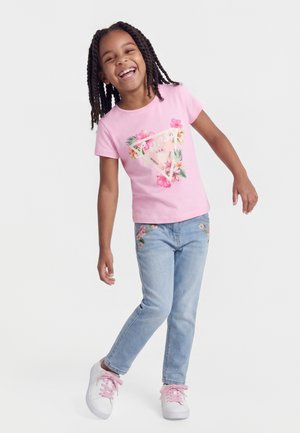 Ragazza giovane con capelli intrecciati che indossa una maglietta rosa con motivi floreali, jeans azzurri ricamati e sneakers bianche con lacci rosa, sorridente e in posa.