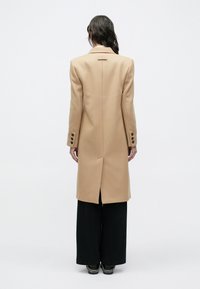 Μπεζ μάλλινο παλτό με κομψή γραμμή, με μία σχισμή στο πίσω μέρος και μαύρα κουμπιά ως διακοσμητικά στοιχεία, σε συνδυασμό με μαύρο παντελόνι φαρδιάς γραμμής.