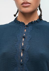 Ναυτικό μπλε μπλούζα με υφή, διαθέτοντας  της ριχτής λαιμόκοψης και δαντέλα κατά μήκος του κέντρου, τονισμένη από διακοσμητικά κουμπιά.