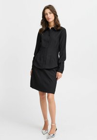 Svart skjorta med knappar och långa ärmar, skräddarsydd passform, i kombination med en matchande svart pennkjol. Silverfärgade högklackade skor kompletterar outfiten.