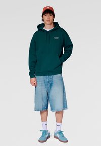 Grüner Kapuzenpullover mit einer Vordertasche, kombiniert mit hellblauen Jeansshorts, türkisfarbenen Turnschuhen und einer korallenfarbenen Kappe. Lässiges Design.