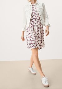 Weiße Jeansjacke über einem ärmellosen lila und weißen gemusterten Kleid mit elastischem Bund. Das Model trägt weiße Sneakers mit pinken Akzenten.