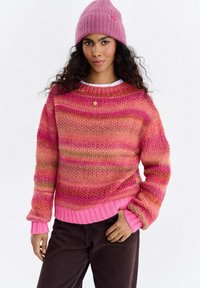 Kobieta z długimi kręconymi włosami, ubrana w różową czapkę z dzianiny, pasek różowo-pomarańczowego swetra, ciemne spodnie oraz złoty naszyjnik z zawieszką.