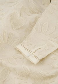 Geborduurde stof met hart- en zonmotieven in een zachte beige kleur, met verhoogde textuur en een knoopmanchetdetails.