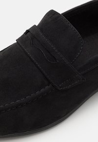 Black suede loafers featuring a stitched trim and an elongated tongue. The upper has a textured finish, with a flat rubber sole for traction.