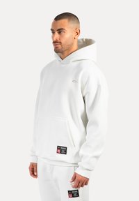Felpa bianca realizzata in tessuto morbido, con tasca frontale, cappuccio con cordino e toppe del marchio sull'orlo. Modello che indossa pantaloni coordinati.