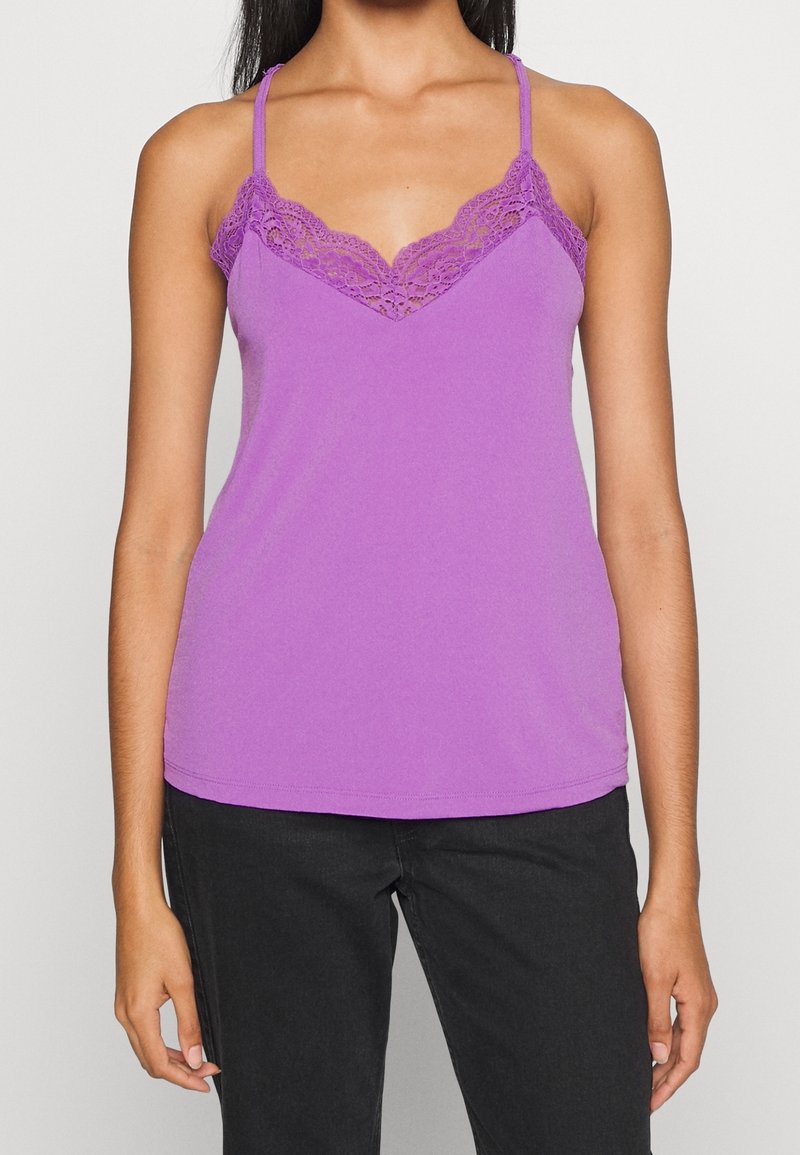 Kobieta w bezrękawnikowej bluzce z fioletowej koronki, wykończonej koronką, w połączeniu z czarnymi spodniami, widoczna od ramion do bioder.