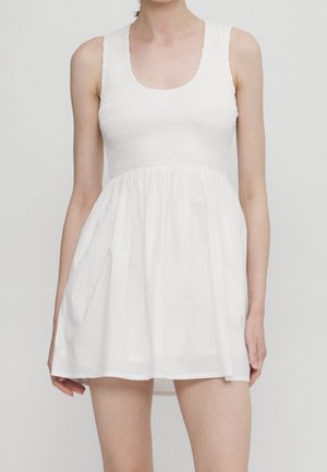 Kobieta w białej sukience bez rękawów z dopasowanym marszczonym gorsem i marszczoną spódnicą na jednolitym szarym tle.