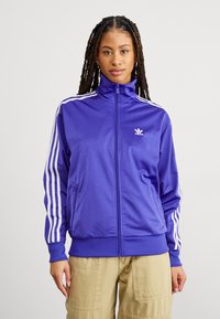 Giacca sportiva viola con colletto alto, zip completa e dettagli a tre strisce bianche sulle maniche. Realizzata in tessuto liscio e lucido.