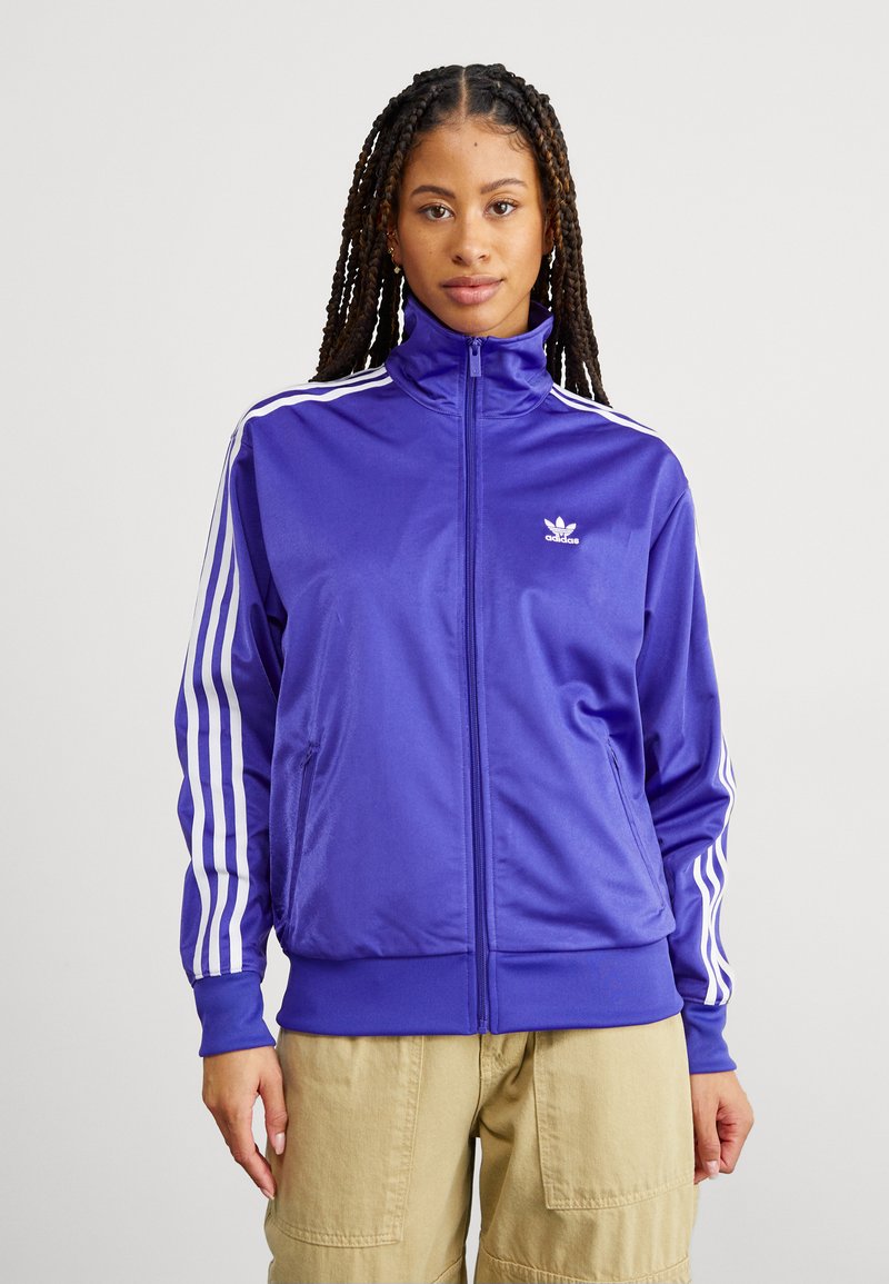 Giacca sportiva viola con colletto alto, zip completa e dettagli a tre strisce bianche sulle maniche. Realizzata in tessuto liscio e lucido.
