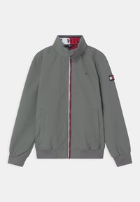 Šedá bomber jacket s vysokým límcem, předním zipem a kontrastním červeným a bílým zipem. Obsahuje elastické manžety a logo nášivku na rukávu.