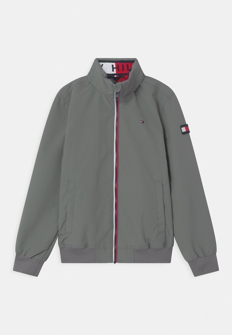 Šedá bomber jacket s vysokým límcem, předním zipem a kontrastním červeným a bílým zipem. Obsahuje elastické manžety a logo nášivku na rukávu.