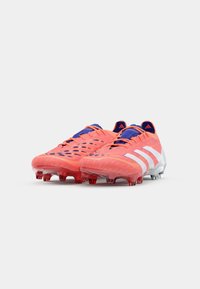 Scarpe da calcio rosse e blu con superficie testurizzata, dotate di strisce bianche, profilo basso e suole trasparenti rosse con tacchetti.