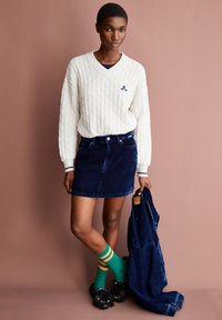 Witte kabelgebreide sweater met V-hals, gecombineerd met een donkere spijkerrok. Groene sokken met gele strepen, zwarte schoenen met franjen.