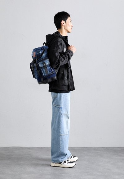 Denim rugzak met een trekkoordsluiting, meerdere ritszakken en een blauwe kleur. Gemaakt met een zwarte jas, zwart shirt en lichtblauwe spijkerbroek.