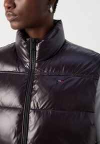 Sjajni, tamnopurpuirani puffer prsluk s visokim kragnom, zatvaranjem na zip, i detaljima loga u crvenoj, bijeloj i plavoj boji. Tekstura na ramenima.