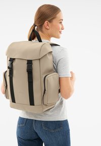 Beige hátizsák felgöngyölíthető tetejű, állítható pántokkal, fekete részletekkel, két oldalsó zsebbel, és sima, matt felülettel.