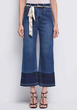 Vaqueros azules anchos y cortos con dobladillo oscuro, cintura alta, cinturón de tela anudado, combinados con sandalias negras de tacón con tiras.