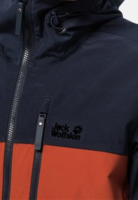 Marinblå och orange vattentät jacka med dragkedja fram, justerbar huva och logotyp på bröstet. Strukturerad tyg med kontrasterande färgpaneler.