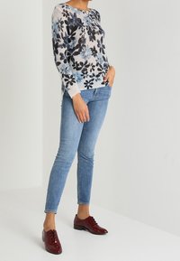 Pull en maille à manches longues avec un motif floral bleu sur fond clair, associé à un jean skinny bleu clair et des chaussures à lacets bordeaux.