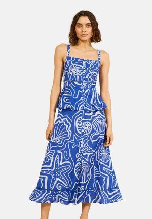 Žena oblečená v modrých šatách bez rukávov s bielymi vzormi morských tvorov, vrátane hviezdic a mušlí, stojaca pred jednoduchým pozadím.