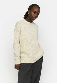 Pulover de culoare crem, cu un model în speckles, guler rotund și manșete ribbate. Are accente cu nasturi pe un umăr. Poartă-l cu pantaloni gri închis.