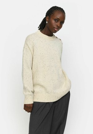 Suéter color crema con un patrón salpicado, cuello redondo y puños acanalados. Presenta botones decorativos en un hombro. Llevado con pantalones gris oscuro.