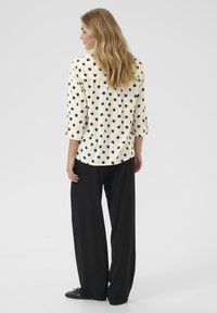 Blusa crema con pois neri, maniche a tre quarti e orlo arrotondato, abbinata a pantaloni neri a gamba larga e scarpe nere.
