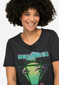 Sort t-shirt med et farverigt grafisk design, der inkluderer teksten "UNIVERSE", palmer og geometriske former i grønne, pink og lilla farver.