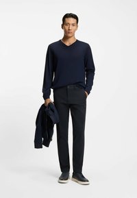 Granatowy sweter z długim rękawem i dekoltem w serek w połączeniu z czarnymi spodniami i granatowymi butami. Jedna ręka trzyma granatową kurtkę. Gładka tekstura, dopasowany krój.