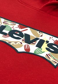 Κόκκινο φούτερ με μια μεγάλη, υφασμάτινη ρίγα γραφικών με παιχνιδιάρικες εικονογραφήσεις φαγητού και τη λέξη "Levi's" σε μαύρα έντονα γράμματα.