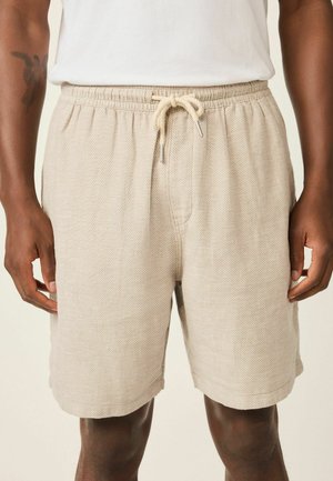 Beeži tekstuuriga lühikesed püksid elastse vöökohaga ja nööriga, mida kannab tumejume inimene vasaku käe tätoveeringuga.