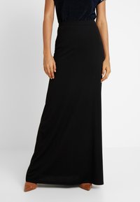 Zwarte, lange, getailleerde maxi-rok met een gladde textuur en een elastische tailleband, gecombineerd met een donkere, sprankelende top en tan-kleurige puntige schoenen.