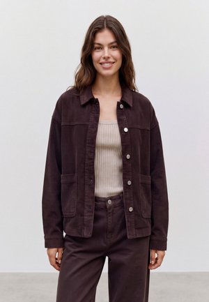 Femme aux longs cheveux ondulés portant une veste en velours côtelé marron foncé, un débardeur côtelé beige et un pantalon marron foncé assorti, souriante à la caméra.