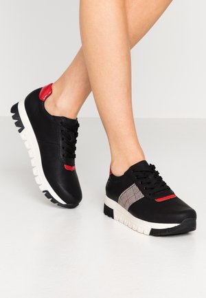 Ben iført sorte sneakers med hvide såler, røde accenter og ternet detalje på siden, mod en hvid baggrund.