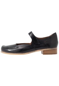 Zapato Mary Jane de cuero negro con punta redonda, correa lateral con cierre de velcro y un tacón bajo de madera apilada. Textura suave.