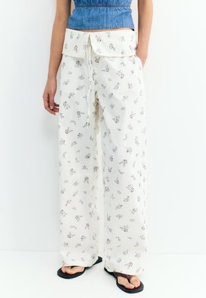 Femeie purtând pantaloni albi cu pantaloni largi, cu un mic imprimeu floral mov, un top albastru fără mâneci și papuci negri, stând pe un fundal alb.