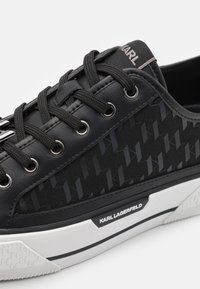 Schwarze Sneakers mit einem Obermaterial aus strukturiertem Stoff, das ein geometrisches Muster aufweist, schwarzen Ösen und einer weißen Gummisohle. Logo an der Seite.