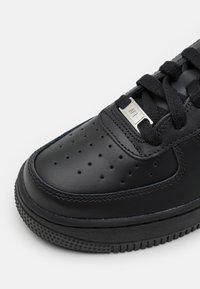 Gros plan d’une basket en cuir noir avec des perforations sur l’orteil, des lacets noirs et une étiquette métallique portant l’inscription "ART" sur les lacets.