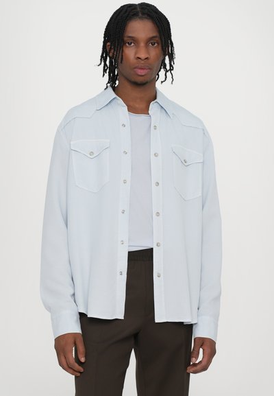 Svetlomodrá džínsová košaľa s dvoma náprsnými vreckami, striebornými cvokmi a voľným strihom, v kombinácii s čiernymi nohavicami s elastickým pásom.