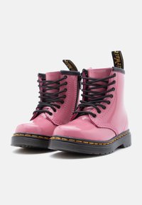 Γυαλιστερά ροζ μποτάκια με κορδόνια, μαύρες σόλες και κίτρινα ράμματα, με ετικέτες με το λογότυπο στο πίσω μέρος, το ένα δίπλα στο άλλο σε λευκό φόντο.